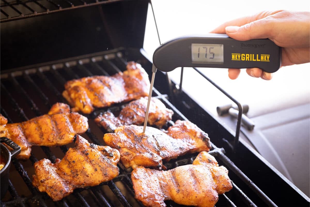 An instant read probe thermometer that reads 175 degrees inserted into a chicken thigh on a grill.