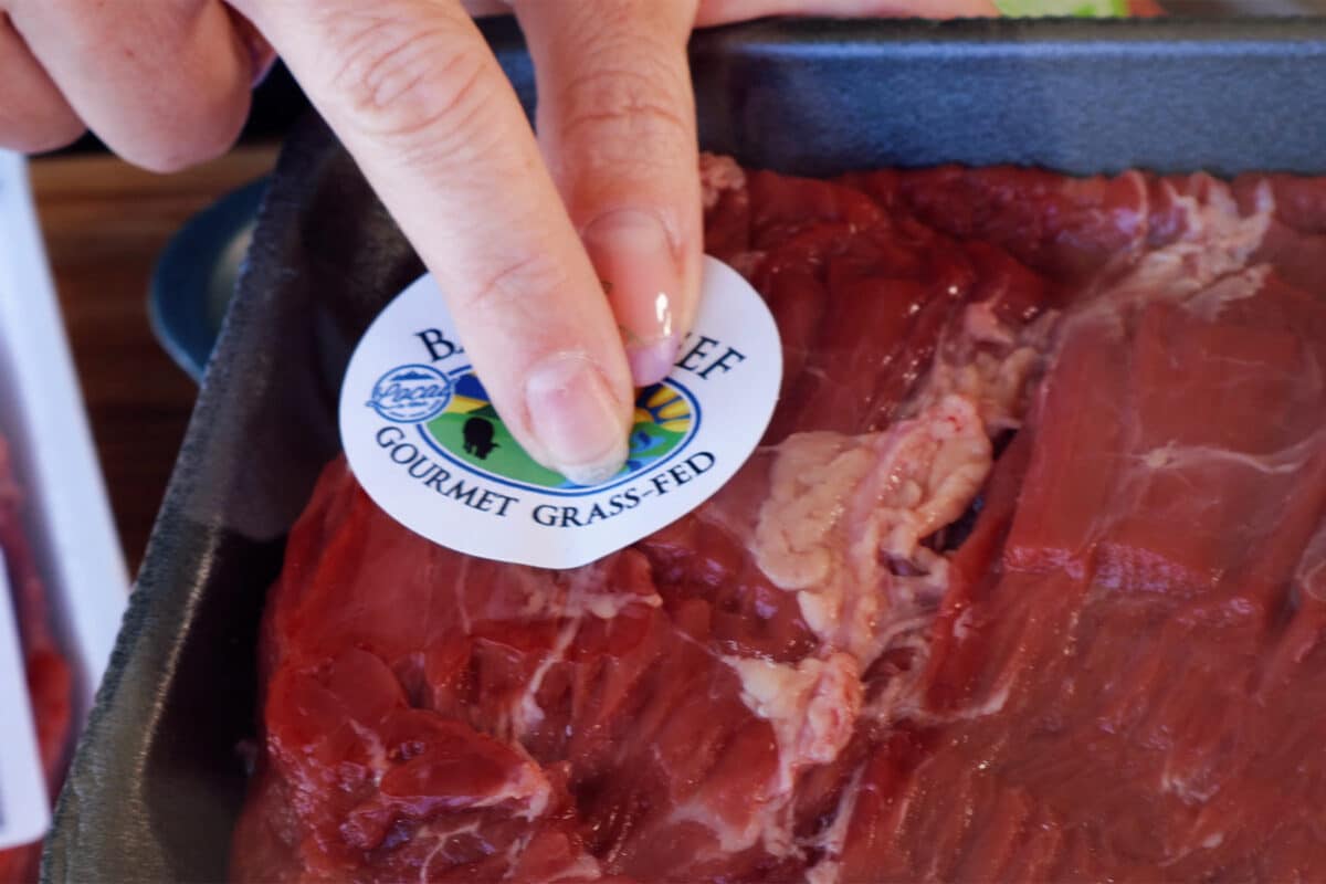 a woman pointing to a sticker on a package of raw meat that says, "Gourmet grass fed".