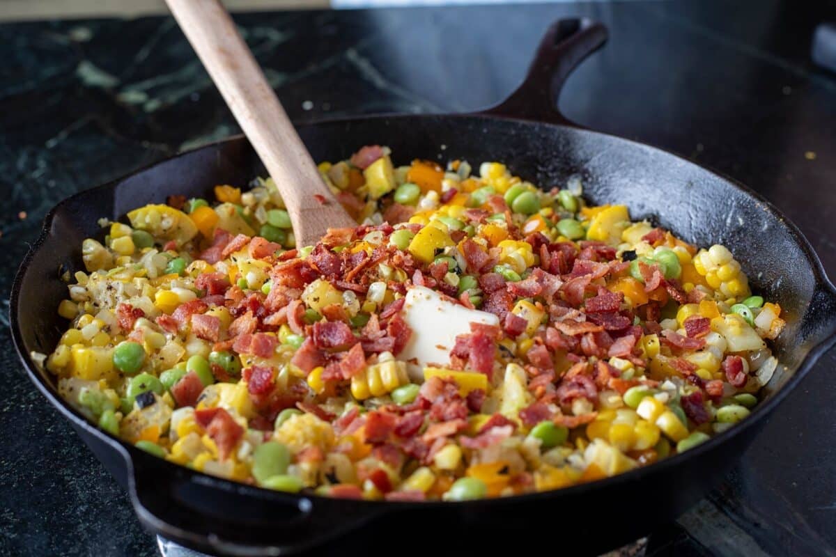 Succotash topped with chopped bacon in a skillet, being stirred with a wooden spoon.