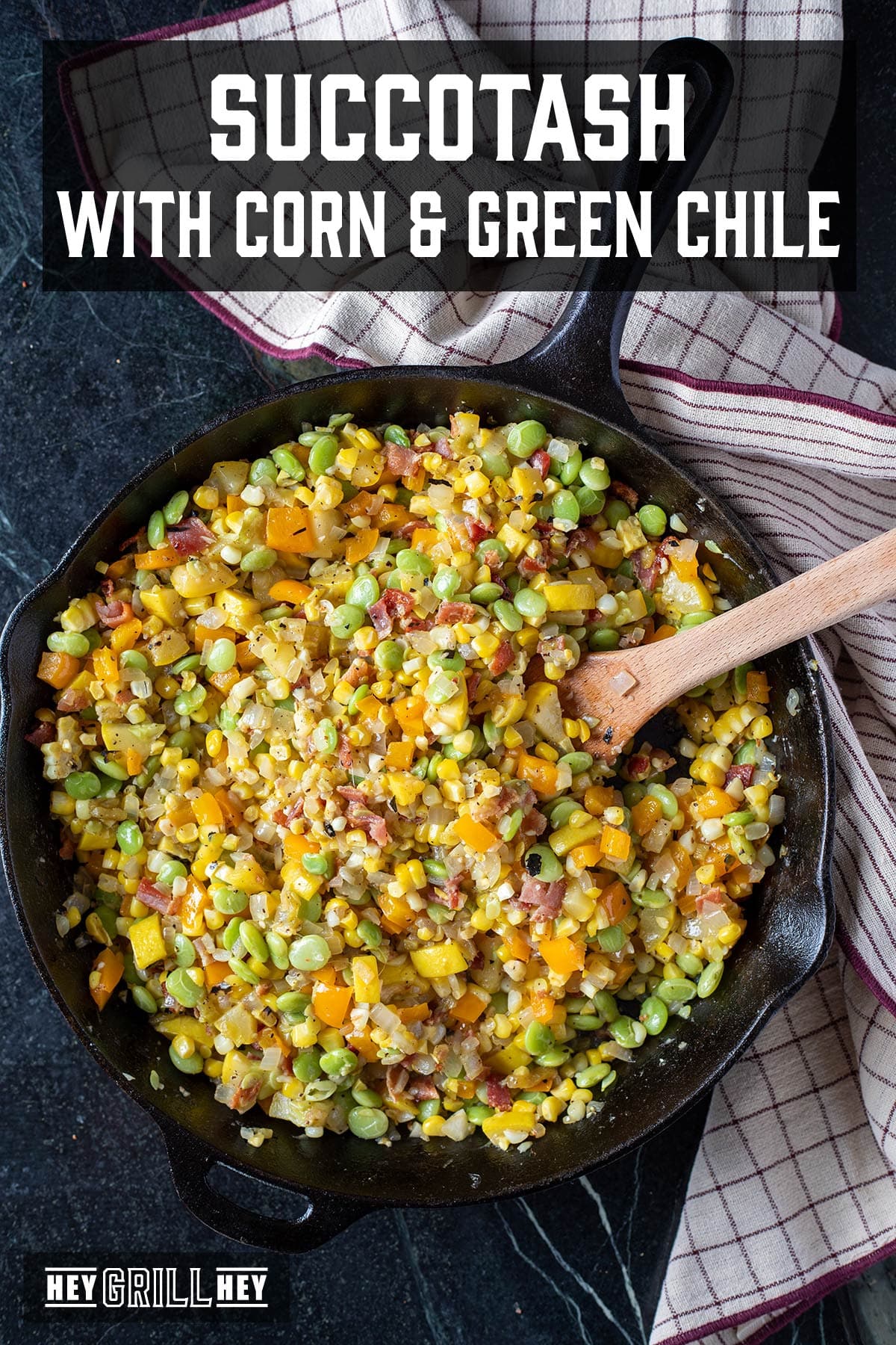 A skillet full of succotash with a wooden spoon resting inside, next to a cloth napkin. The text overlay and the top reads "Succotash with Corn and Green Chile," and the small one at the bottom reads "Hey Grill Hey."