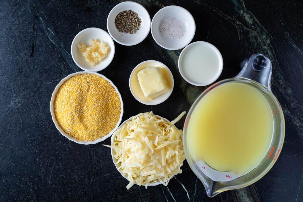 Chicken broth, shredded cheese, cornmeal, and other ingredients for grits in containers on a countertop.