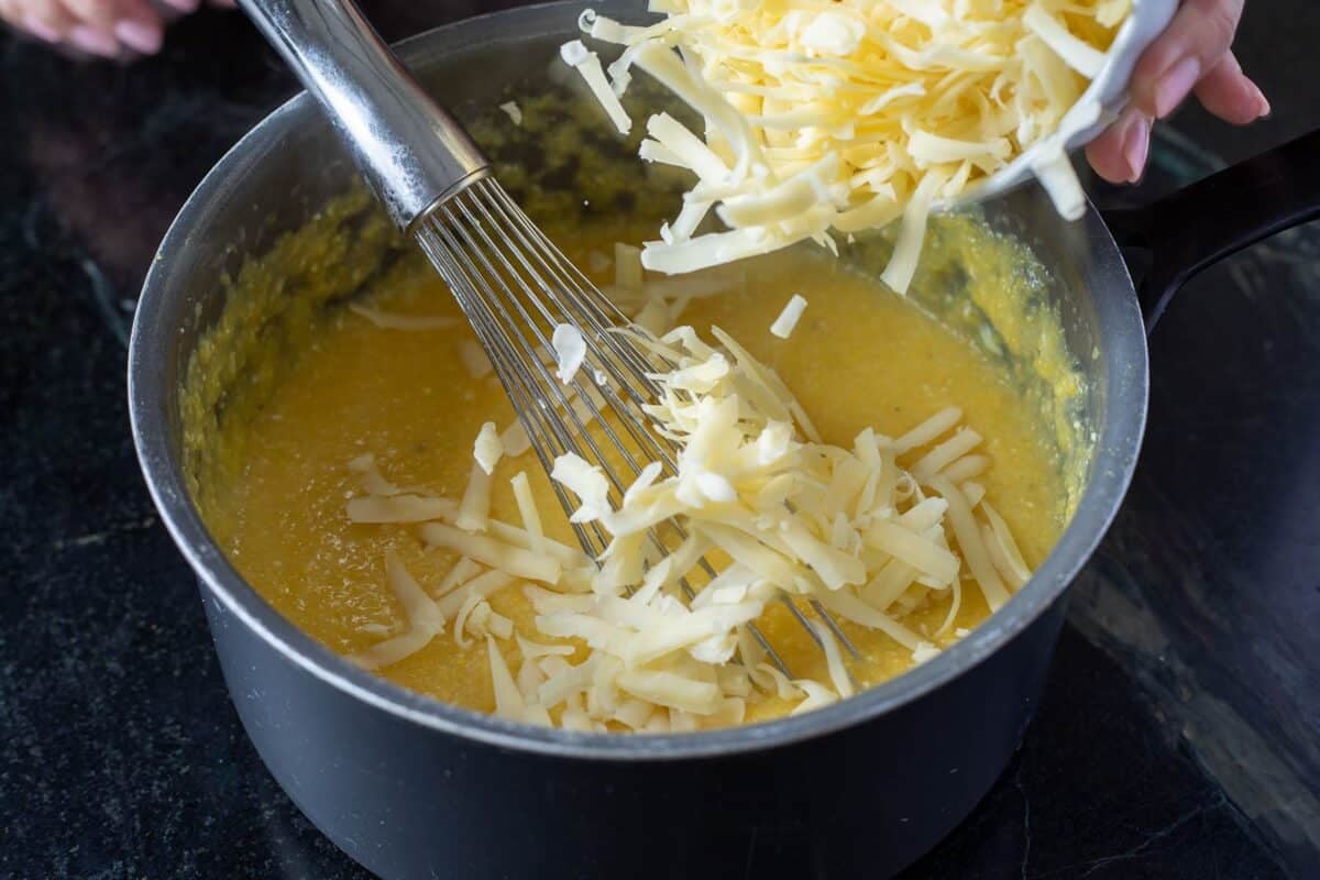 Shredded cheese being sprinkled into a mixture being whisked in a pot.