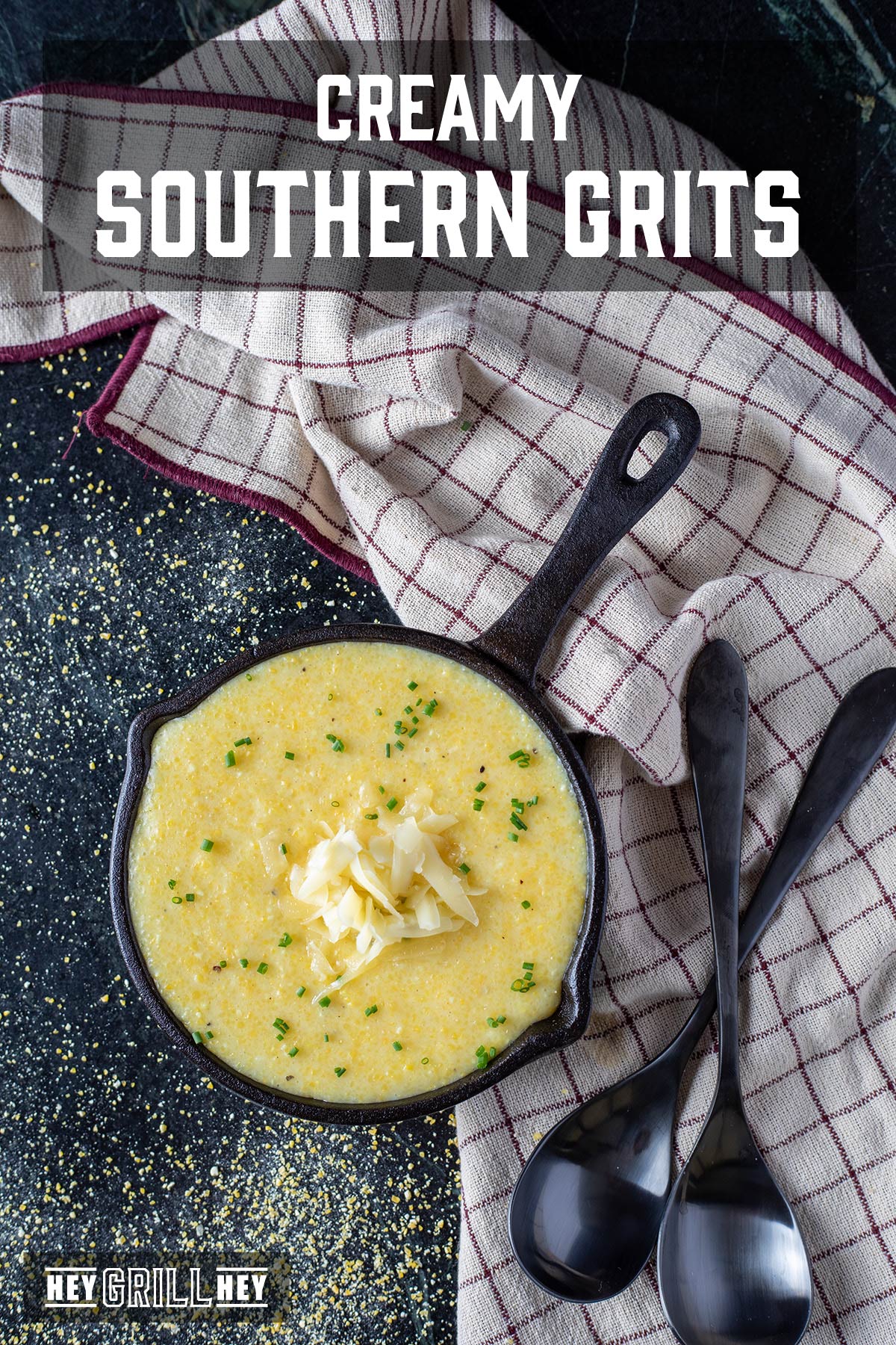 A cast iron pan filled with grits topped with shredded cheese. There's a tablecloth next to it with 2 spoons on it. The text overlay reads "Creamy Southern Grits" at the top, and "Hey Grill Hey" at the bottom.