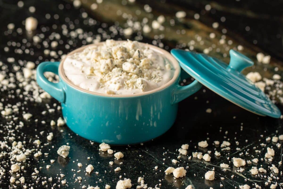 Blue cheese dressing in a blue pot on a black surface sprinkled with blue cheese crumbles.