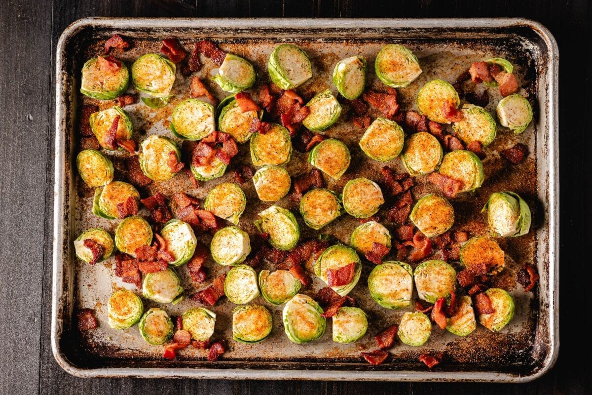 Brussels sprouts halves and chopped bacon on a metal baking sheet coated with BBQ seasoning.