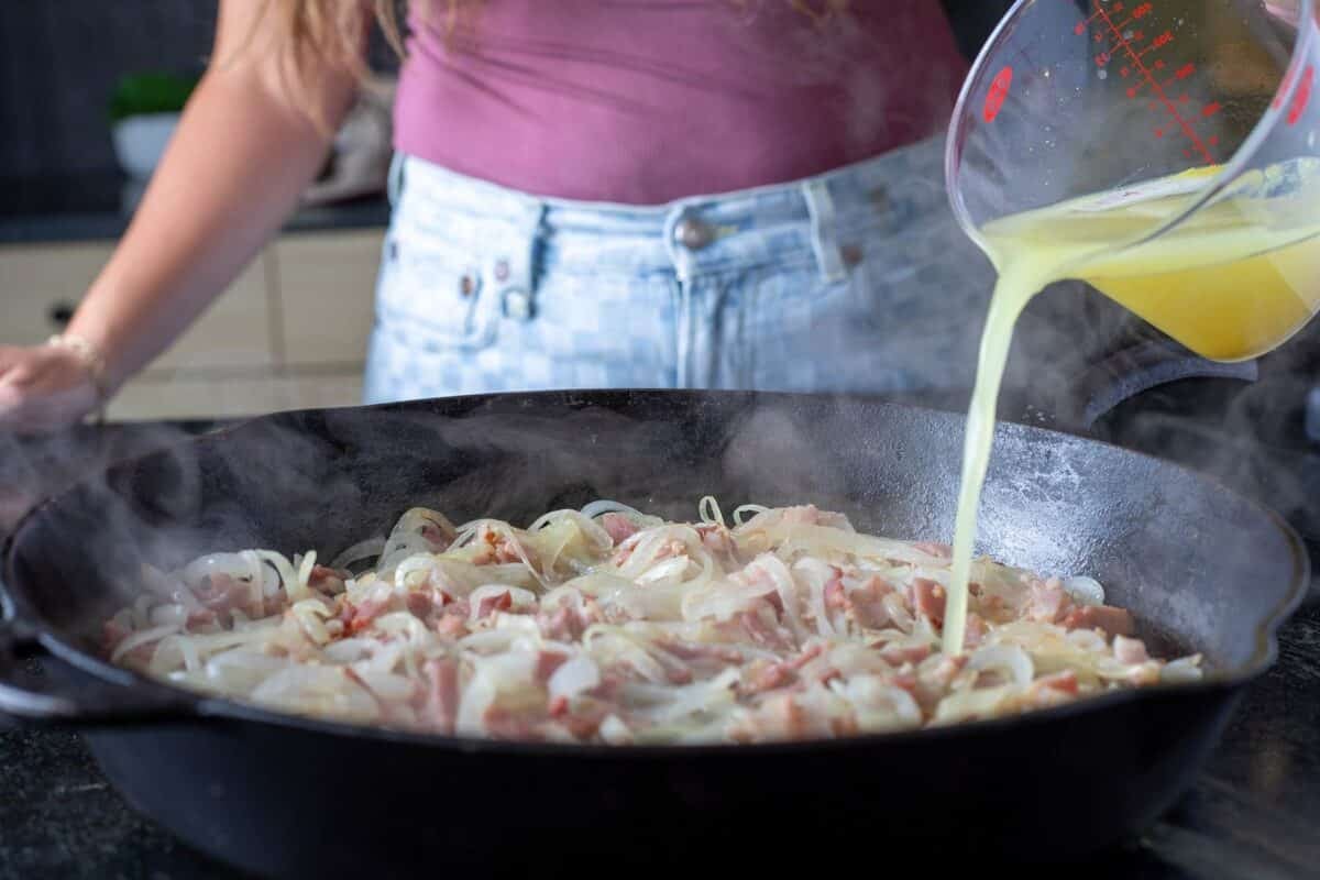 Bacon and onions in a skillet, with a cup of broth being poured into it.