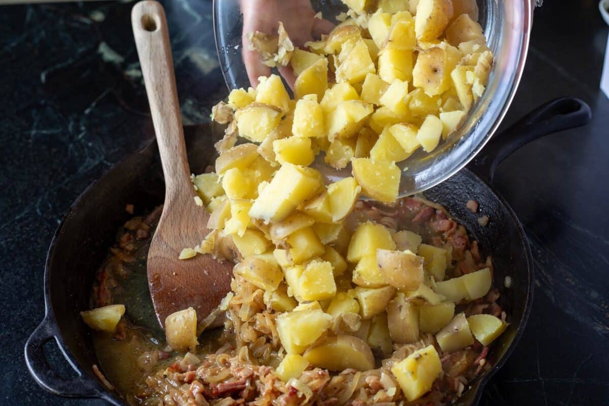 A bowl of potato chunks being poured into a skillet of onions.