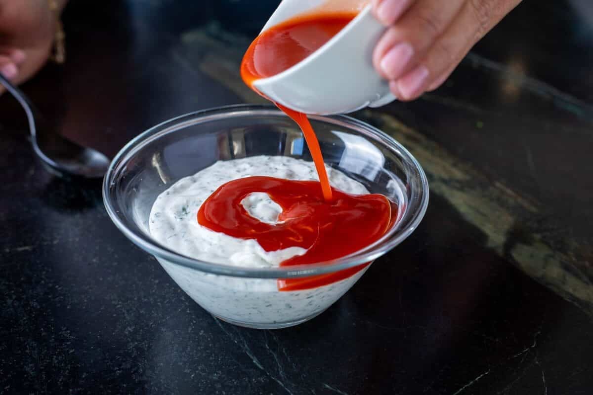 Buffalo sauce being poured from a white bowl into a clear bowl full of ranch dressing.