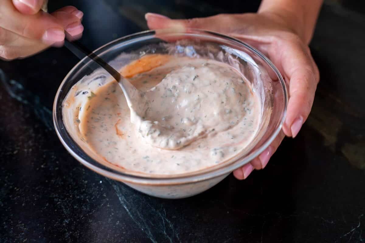 A hand holding a clear bowl full of sauce while a another hand stirs it with a spoon.