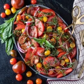 Tomato salad on a serving platter next to loose tomatoes, basil garnish, and serving tongs.