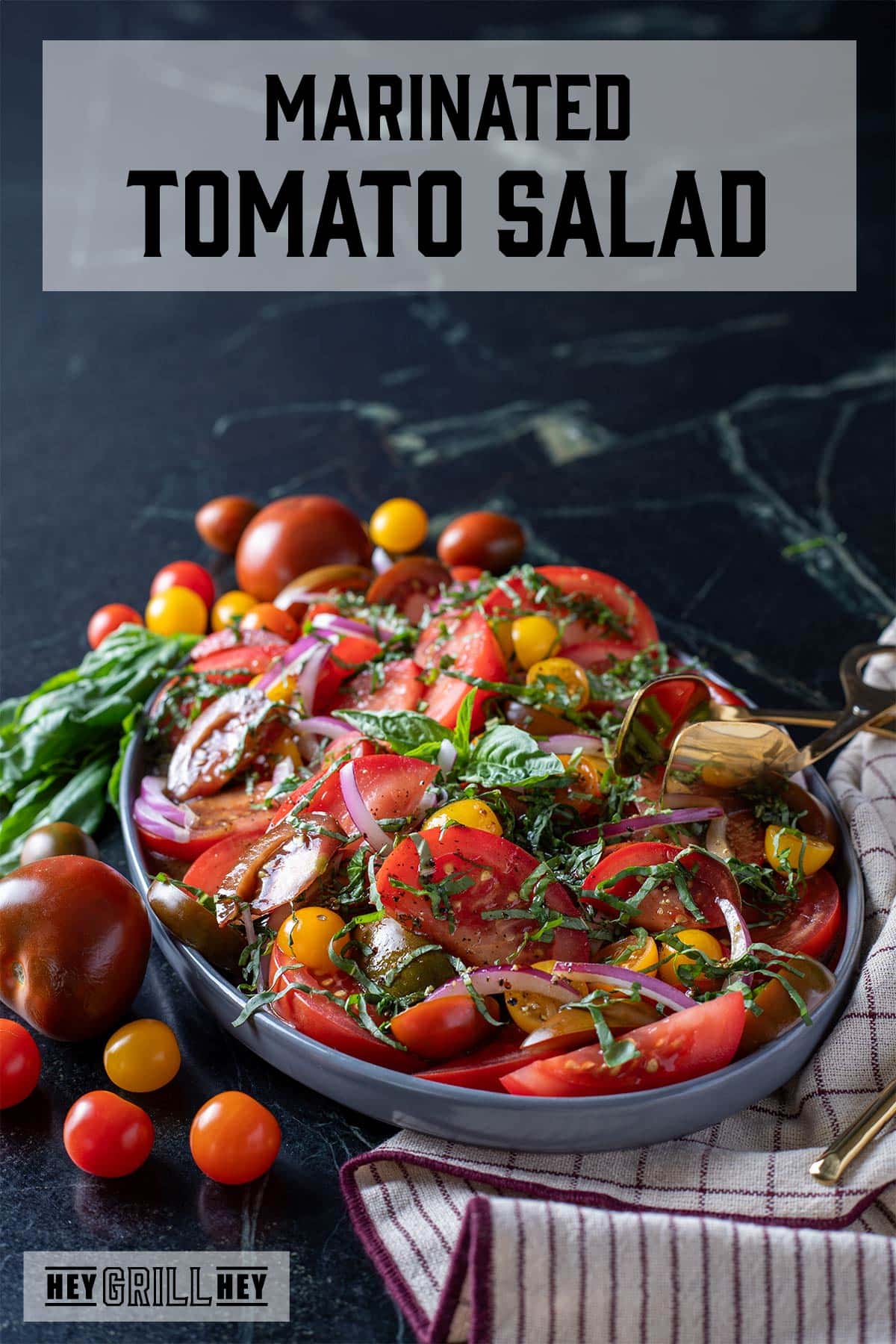 Tomato salad on a serving platter, garnished with whole cherry tomatoes and basil leaves. The text overlay and the top reads "Marinated Tomato Salad," and the small one at the bottom reads "Hey Grill Hey."