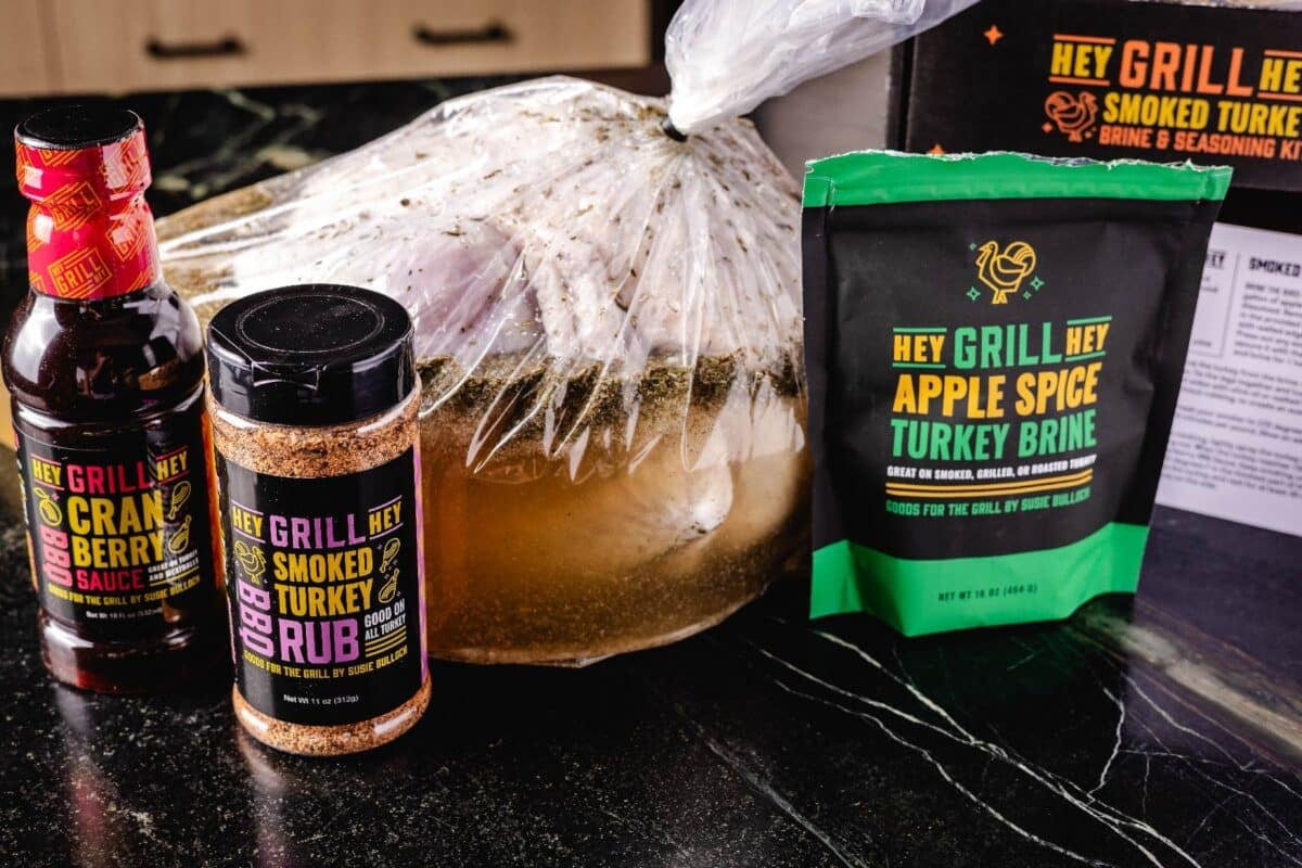 A turkey in a brining bag on a counter next to a bottle of rub and bottle of sauce.