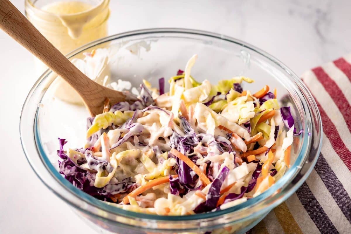 Homemade coleslaw in a glass bowl being stirred with a wooden spoon.