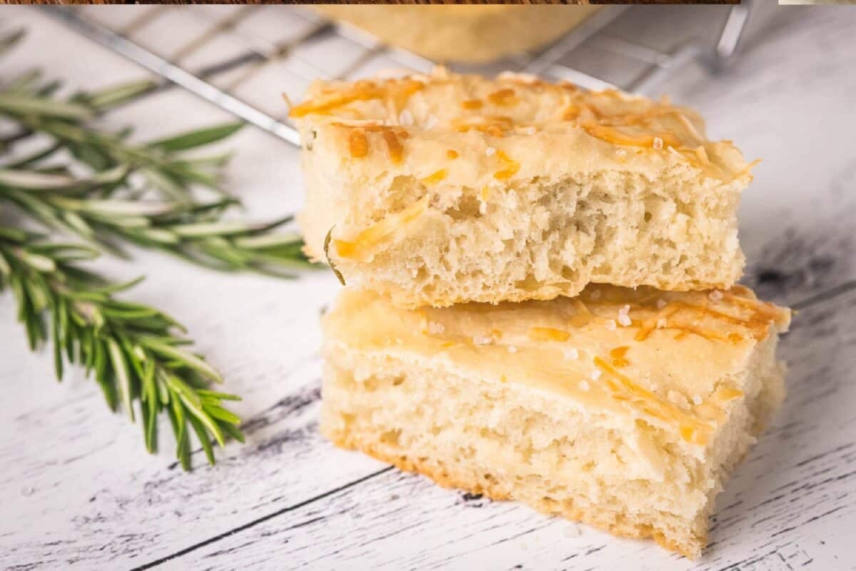 Rosemary focaccia bread slices on a white tablecloth next to fresh rosemary sprigs.