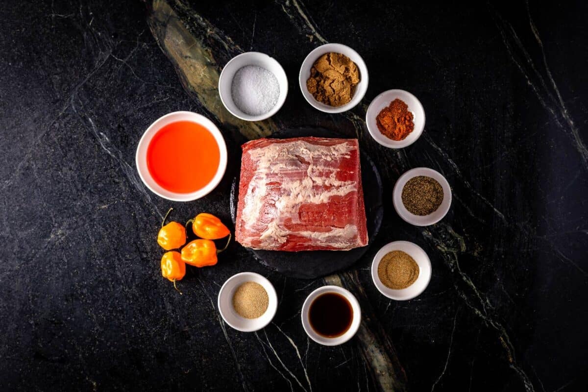 Raw beef roast and chilis on a countertop surrounded by glass bowls full of raw ingredients.