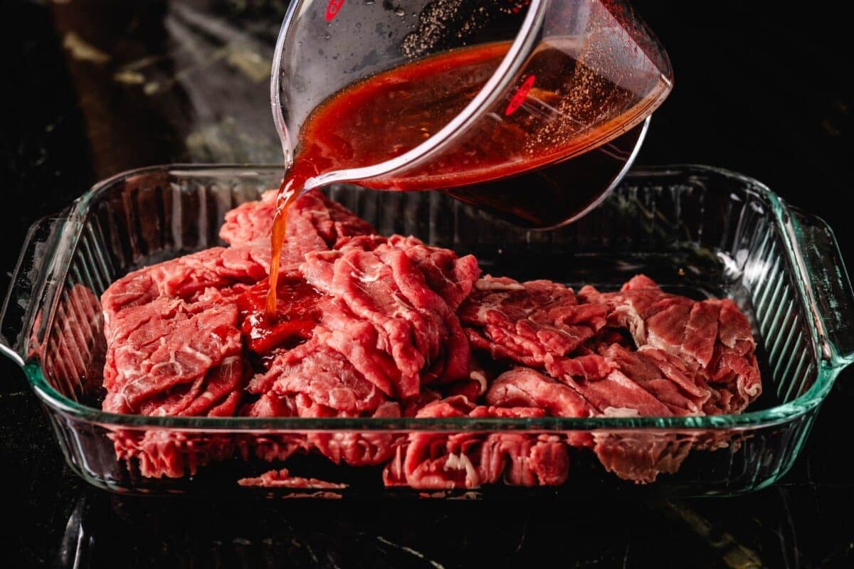 A cup of marinade being poured onto beef slices in a baking dish.