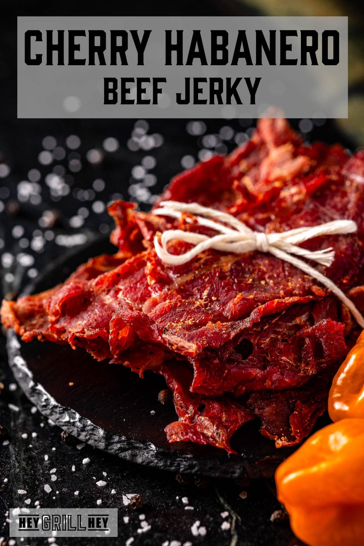 Jerky tied with butcher's twine sitting on a black platter next to habanero peppers. The text overlay reads "Cherry Habanero Beef Jerky" at the top and "Hey Grill Hey" at the bottom.