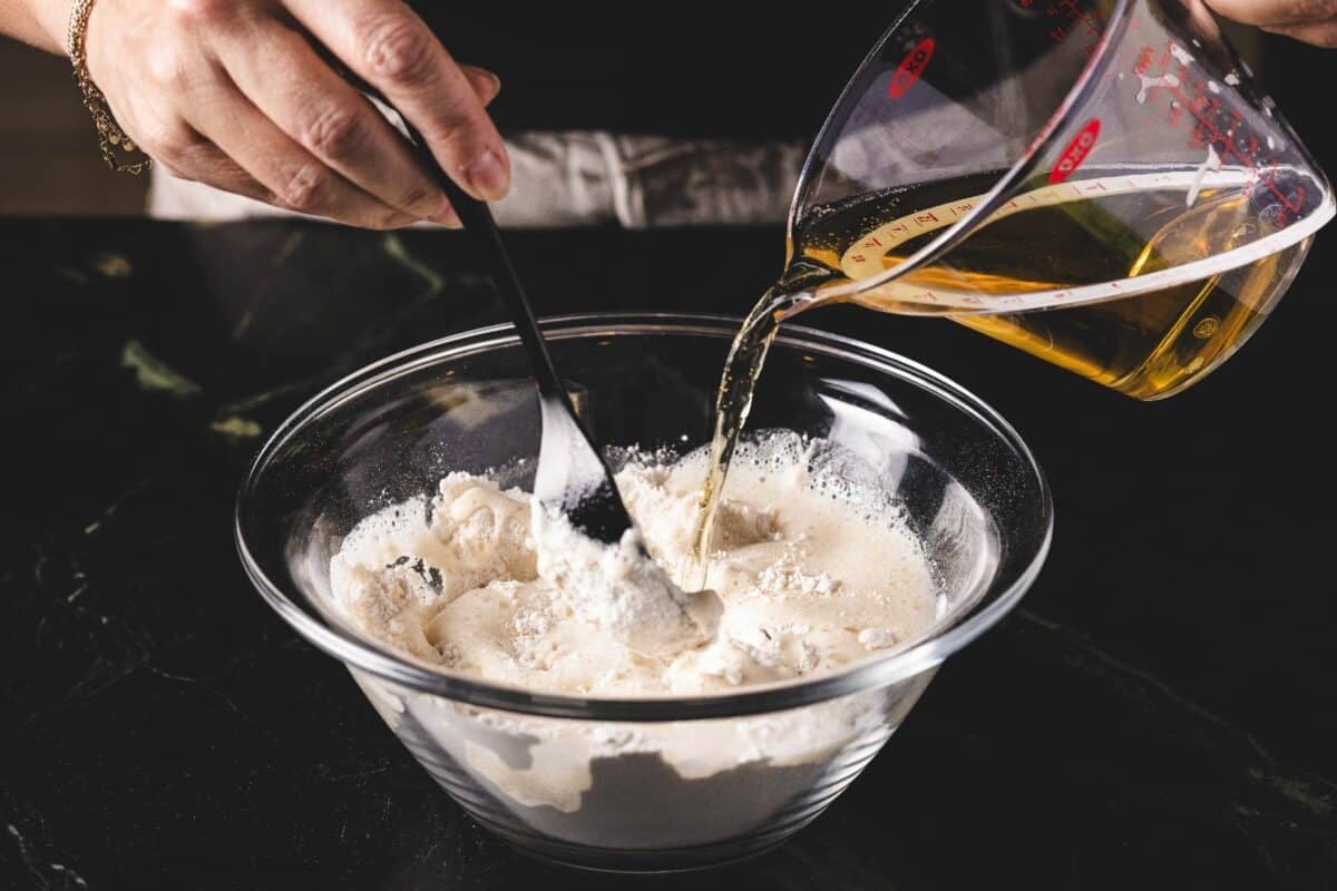 Batter in a glass bowl being mixed with a spoon while beer is poured in from a cup.