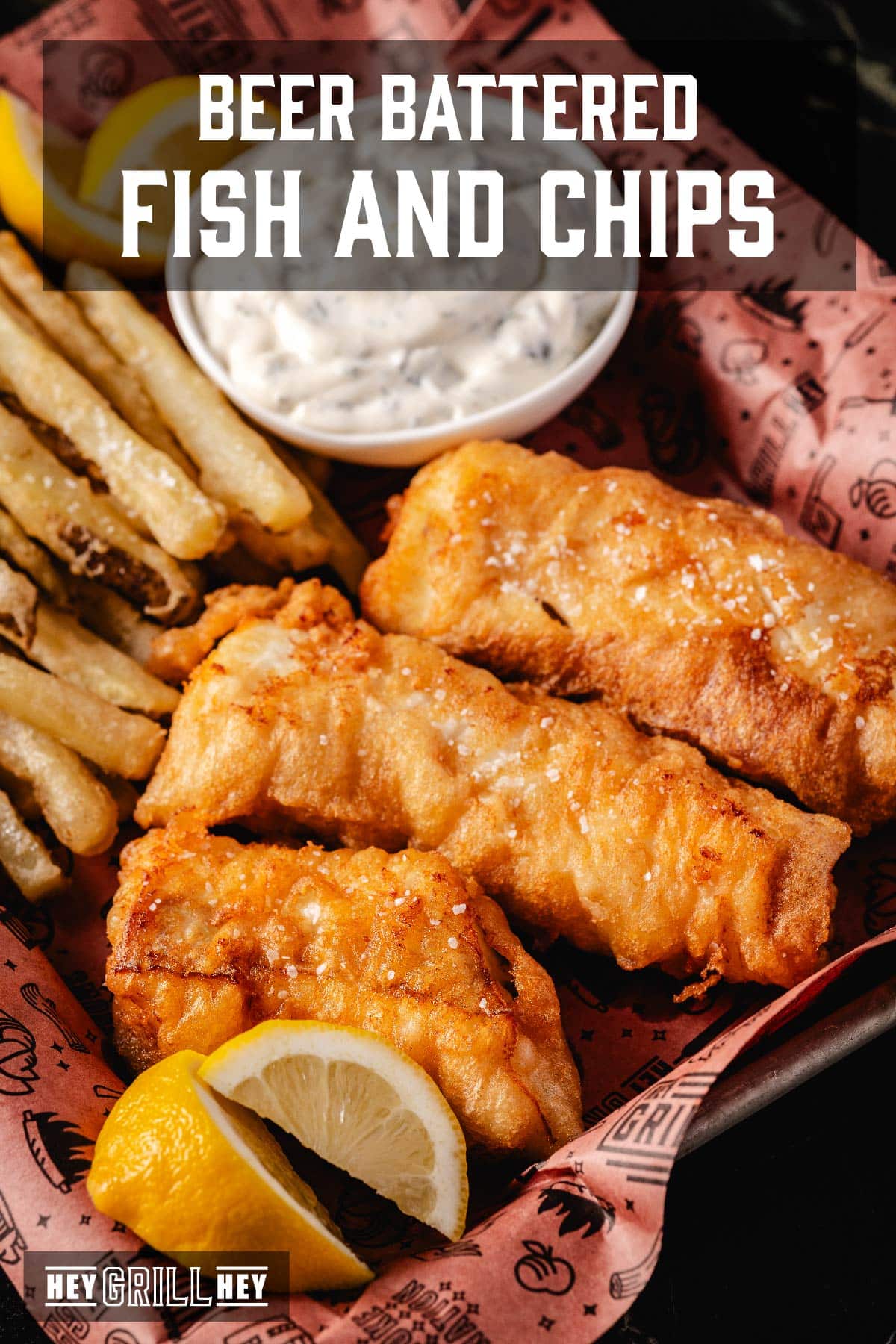 Fried fish fillets, French fries, and a cup of tartar sauce in a paper-lined basket. The text overlay reads "Beer Battered Fish and Chips" at the top, and "Hey Grill Hey" at the bottom.