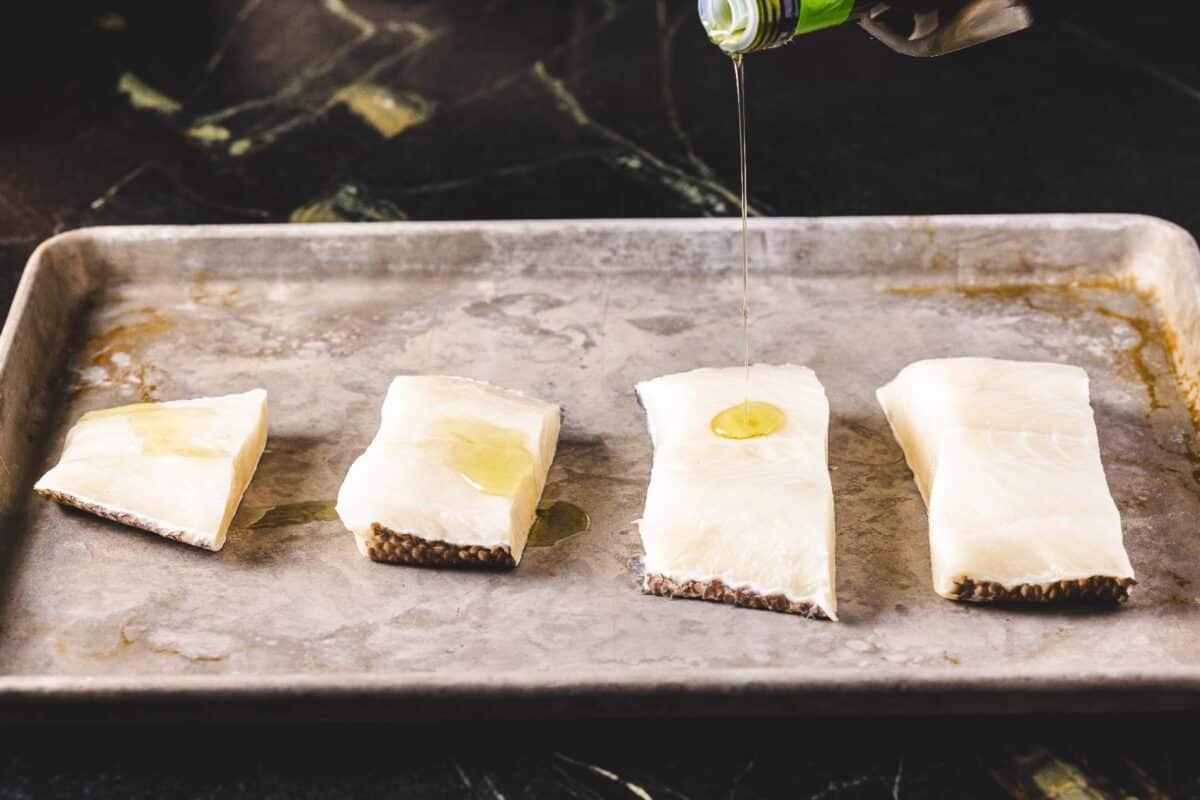 Fish fillets on a baking sheet being drizzled with oil.