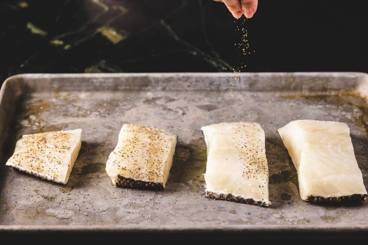 Fish fillets on a baking sheet being sprinkled with salt and pepper.