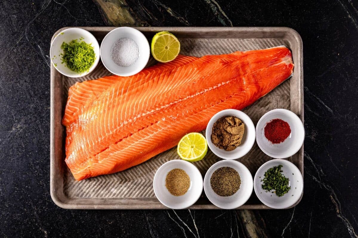 A raw fish fillet on a baking sheet with glass bowls of seasoning ingredients.