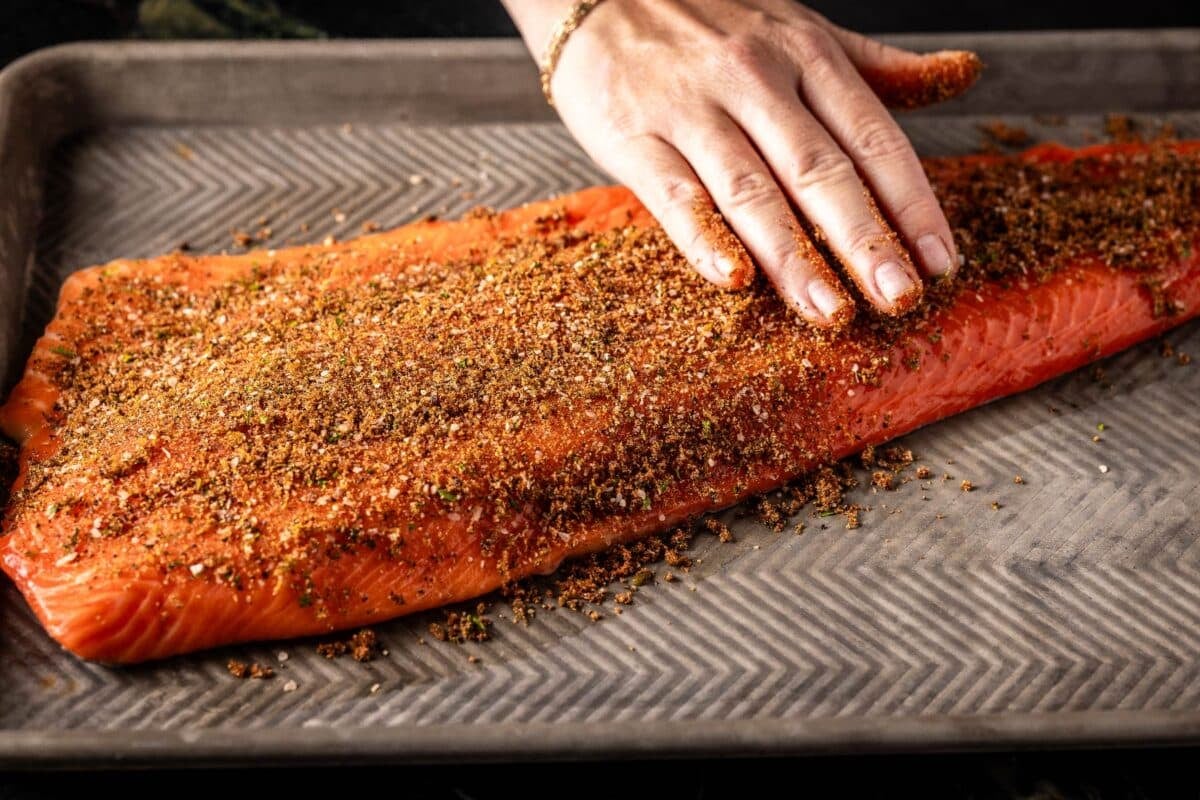 A hand patting seasoning into a fish fillet on a baking sheet.