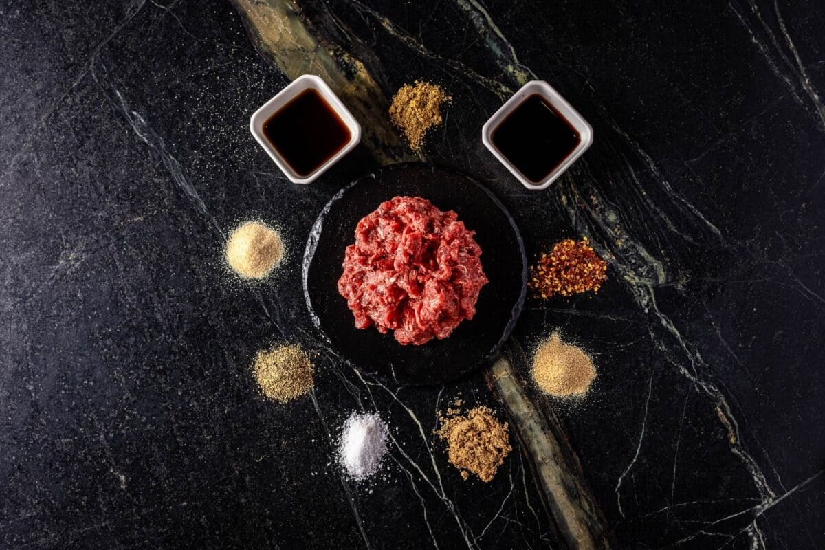 Worcestershire and soy sauces in glass bowls, surrounded by raw seasoning ingredients on a countertop.