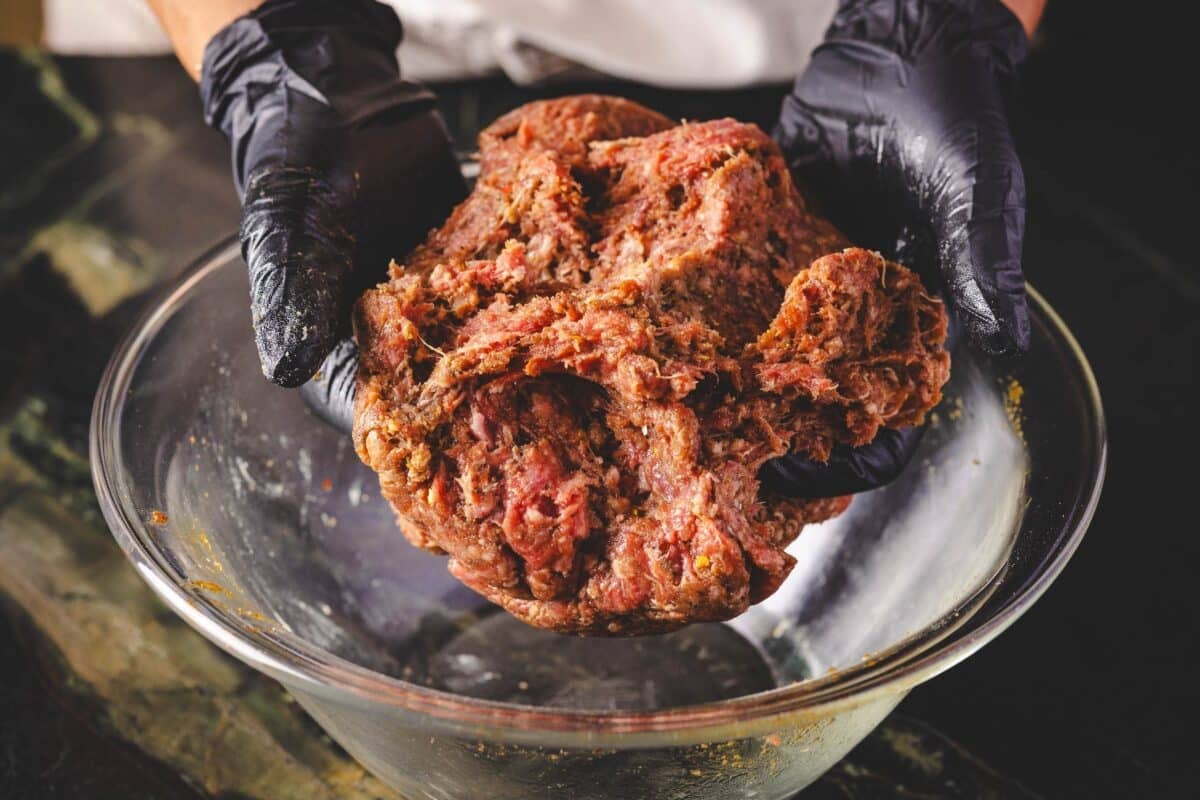 Ground meat being placed in a glass bowl by gloved hands.