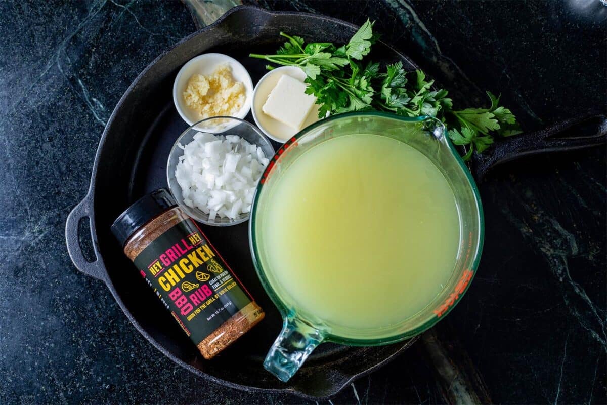 A cast iron skillet on a countertop containing a cup of chicken stock and various herbs and seasonings.