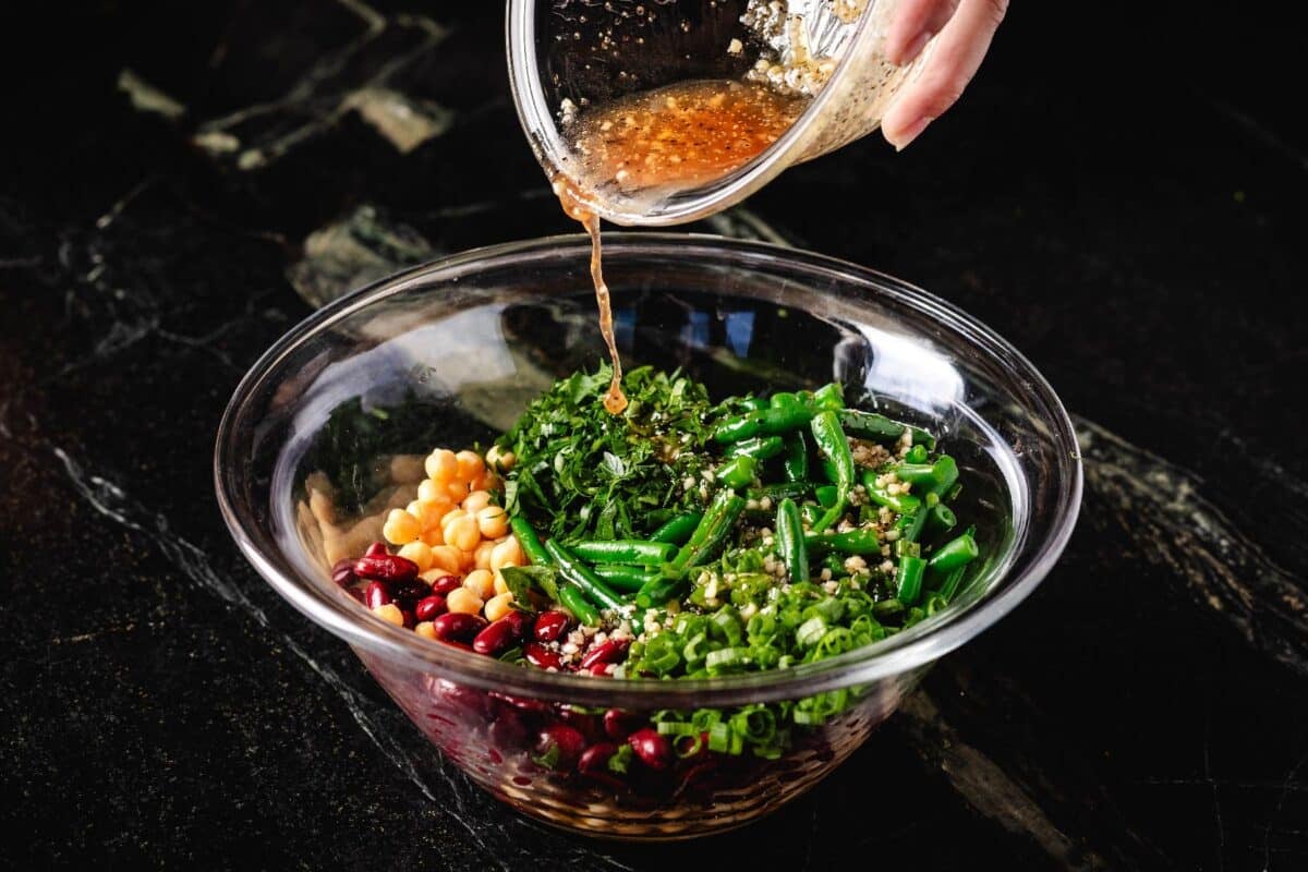 Dressing being poured from a bowl into a larger bowl filled with green, kidney, and garbanzo beans.