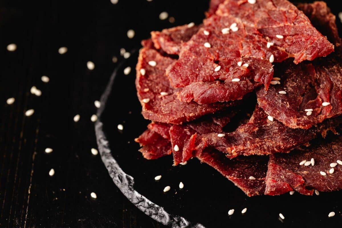 Slices of teriyaki flavored dehydrated beef on a black plate sprinkles with sesame seeds.