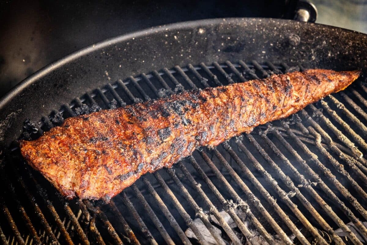 A skirt steak sitting on the grates of a charcoal grill.