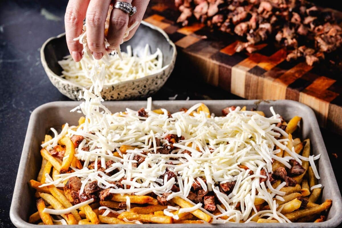 A baking sheet of fries topped with carne asada being sprinkled with shredded cheese.