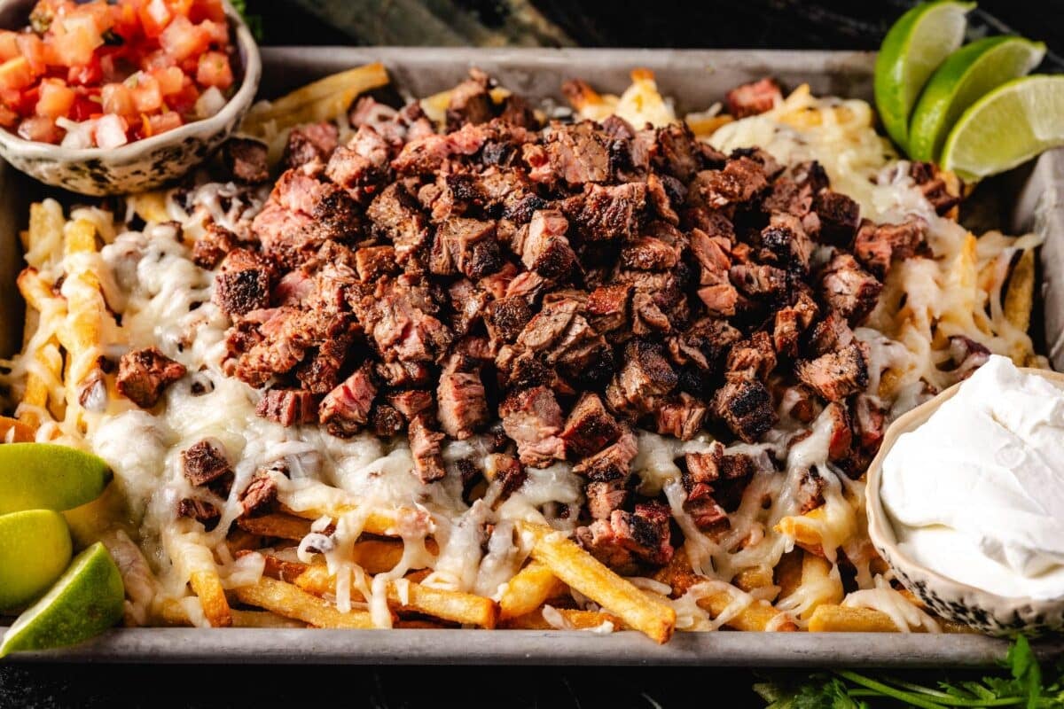 A sheet pan of fries topped with cheese and carne asada, and cuts with sour cream and diced tomatoes.