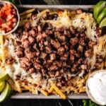 A sheet pan of fries topped with cheese and carne asada, and cuts with sour cream and diced tomatoes.