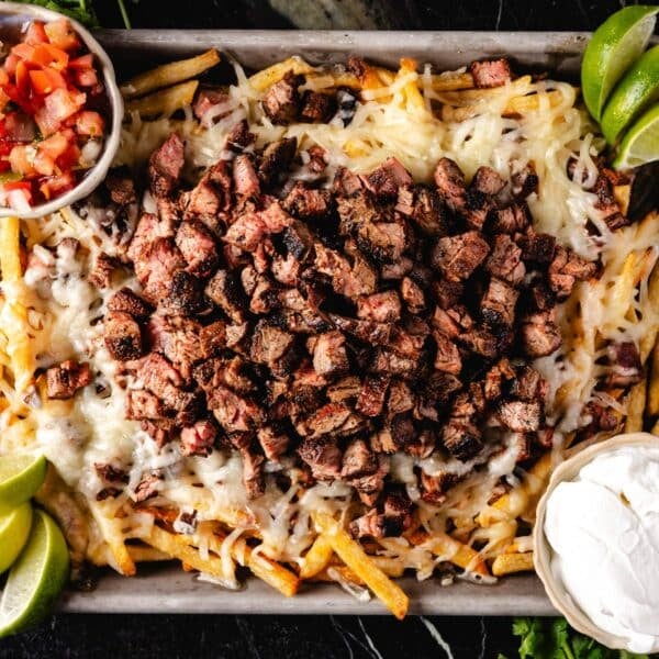 A sheet pan of fries topped with cheese and carne asada, and cuts with sour cream and diced tomatoes.