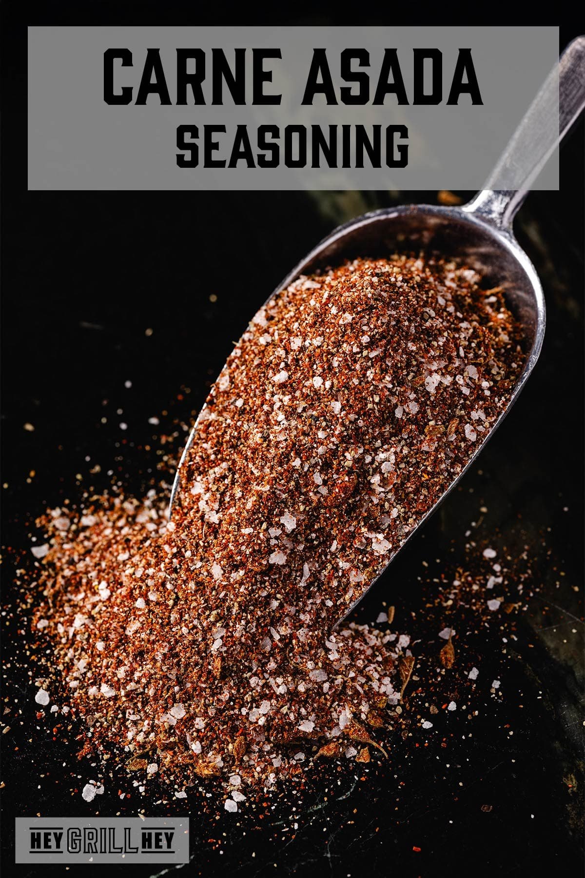 A scoop overflowing with seasoning on a black surface. The text overlay reads "Carne Asada Seasoning" at the top, and "Hey Grill Hey" at the bottom.