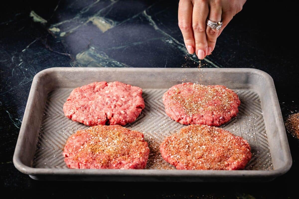 Four raw burger patties on a baking sheet being sprinkled with seasoning.