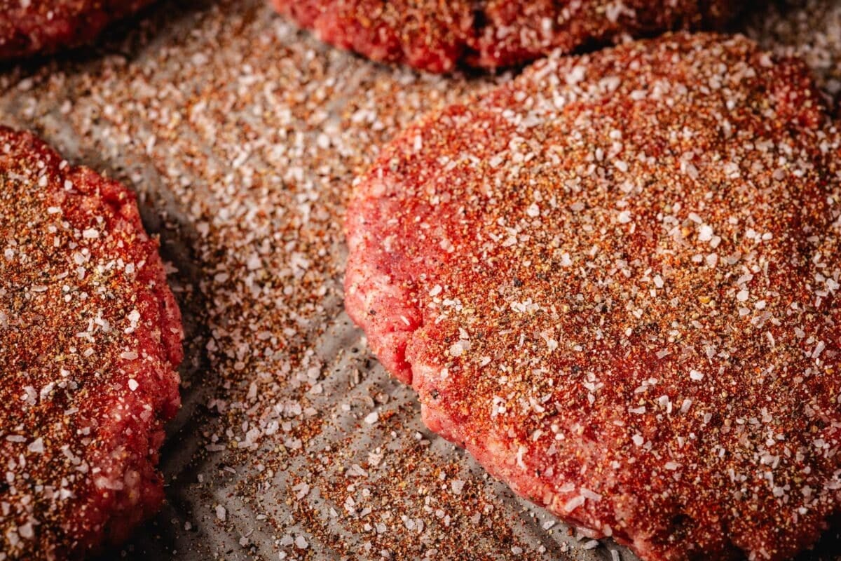 Raw burger patties on a baking sheet dusted with seasoning.