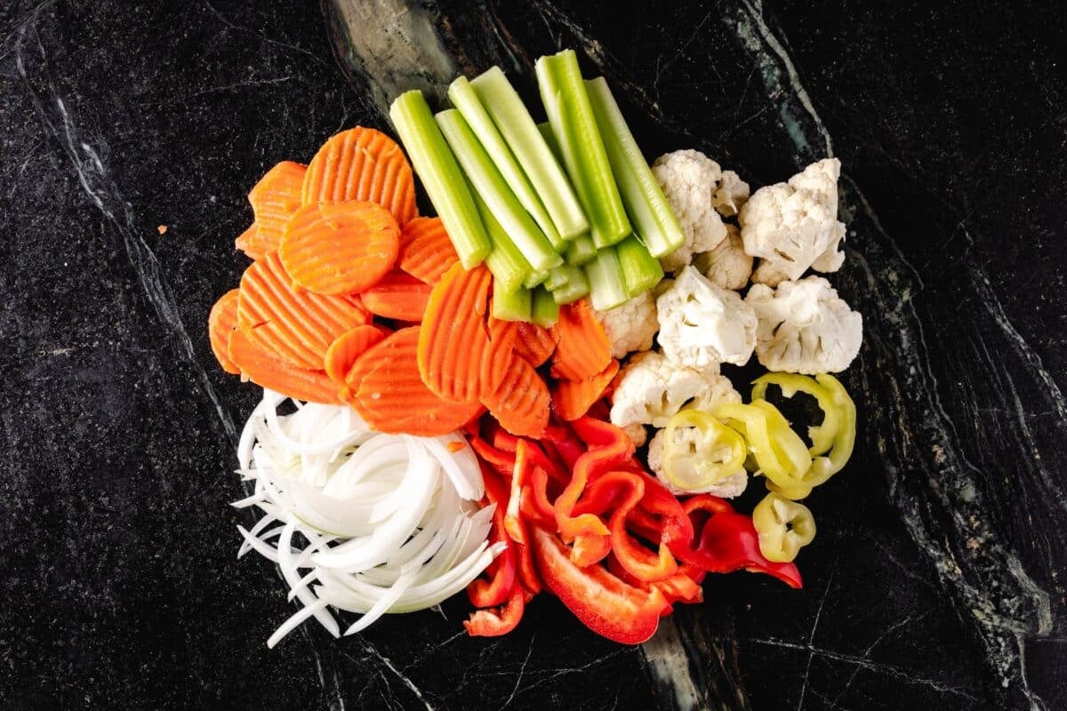 Sliced carrots, celery, cauliflower, peppers, and onions in a pile on a granite counter.