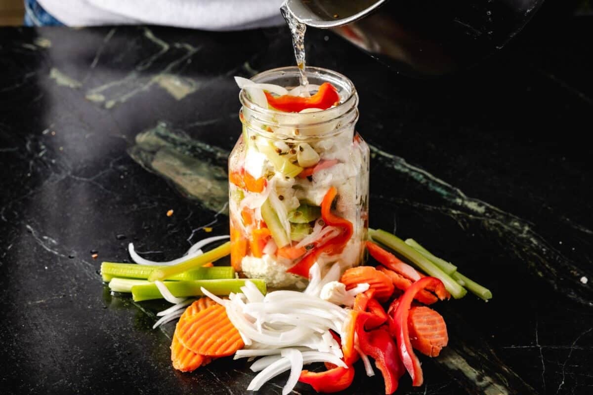 A glass jar filled with relish, with sliced onions, carrots, and peppers laying in front of it.