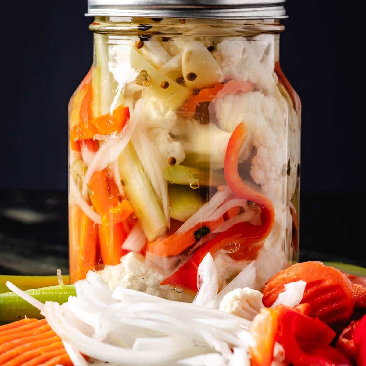 A glass jar filled with giardiniera, with sliced onions, carrots, and peppers laying in front of it.