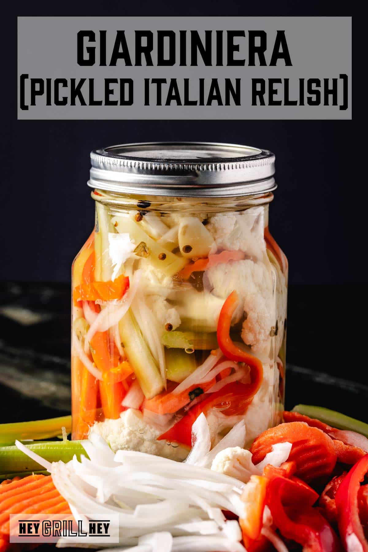 A glass jar filled with relish, with sliced onions, carrots, and peppers laying in front of it. The text overlay reads "Giardiniera (Pickled Italian Relish)" at the top, and "Hey Grill Hey" at the bottom.