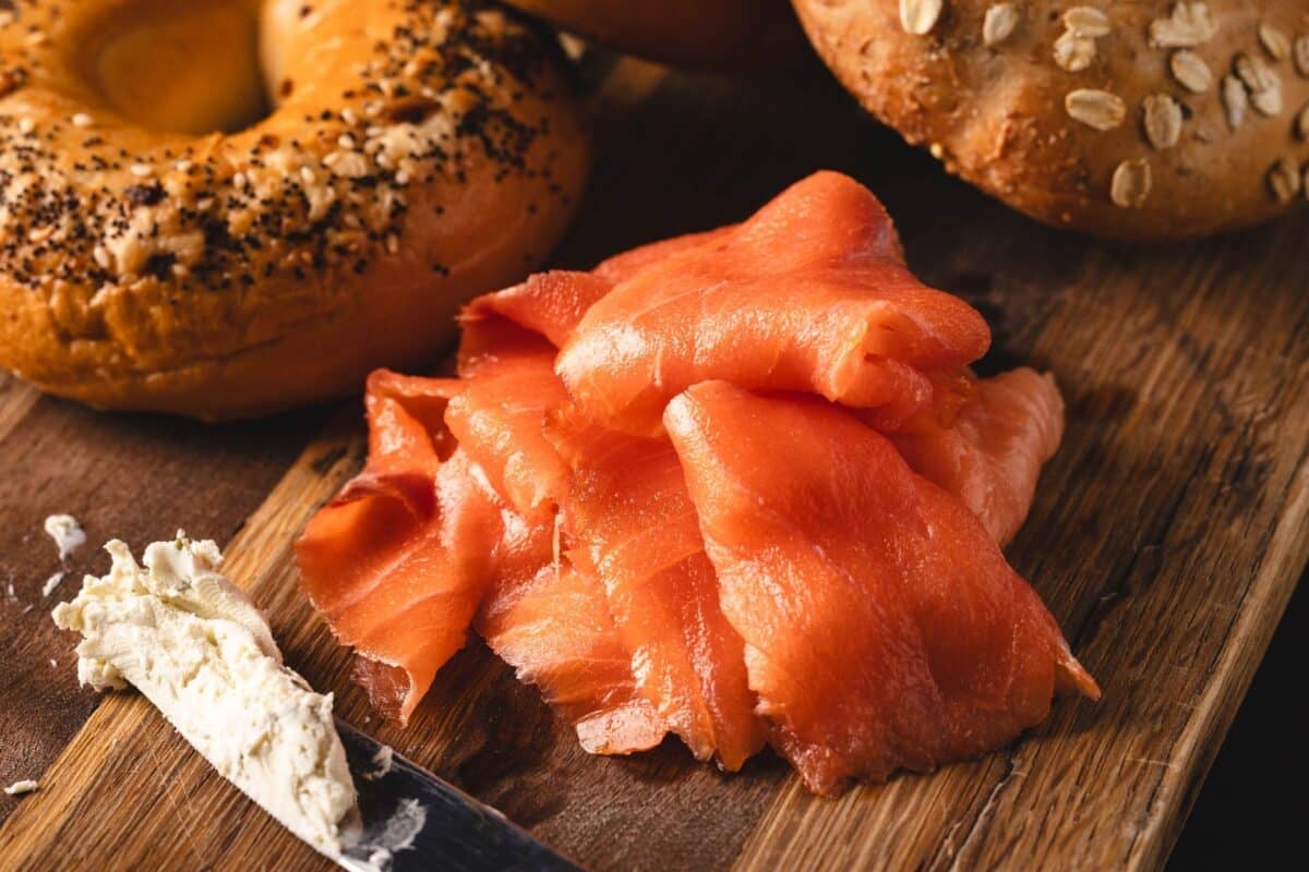 Sliced, cold smoked salmon on a cutting board with bagels, and a knife with cream cheese on it.