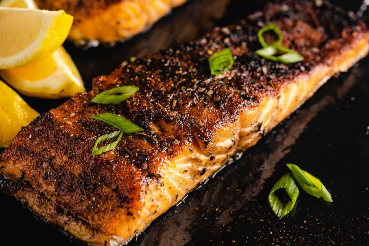 A blackened salmon fillet on a black surface next to lemon wedges.