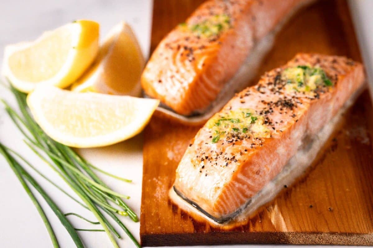 Salmon fillets on a cedar plank next to lemon wedges and herbs.