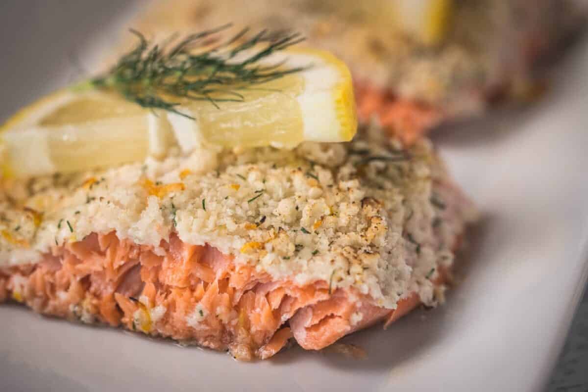 An herb crusted salmon fillet topped with a lemon slice and dill garnish on a white plate.