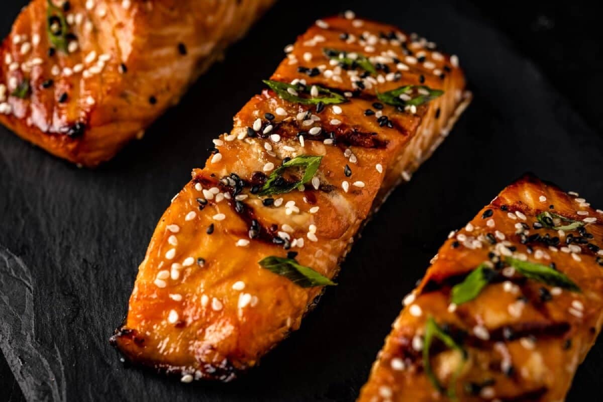 Grilled fish fillets topped with sesame seeds and chives on a black platter.