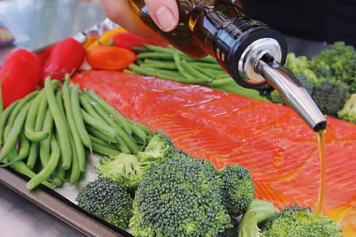 Oil being drizzled from a bottle onto a fish fillet and veggies on a sheet pan.